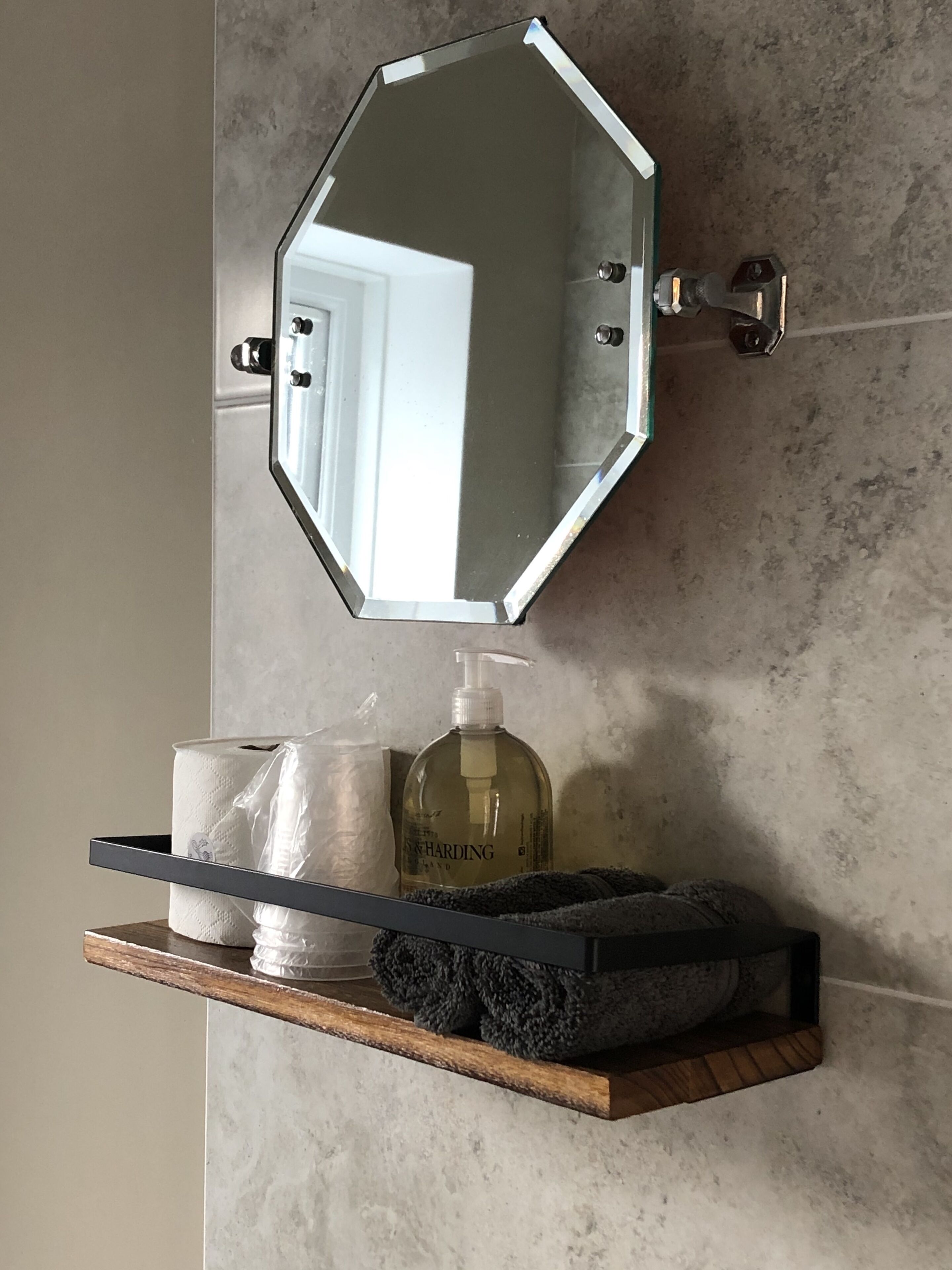 Et badeværelse med et hexagonalt spejl, en træhylde med toiletartikler og en marmorvæg.