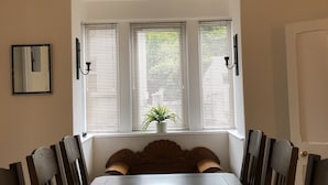 A dining room with a long table, three windows with blinds, a potted plant, and a hanging light fixture. - Seacot Cottage in the heart of the Highlands (Clachnaharry)