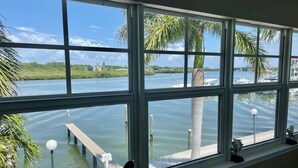 A view through a large window with multiple panes, overlooking a calm body of water, a dock, and a palm tree.