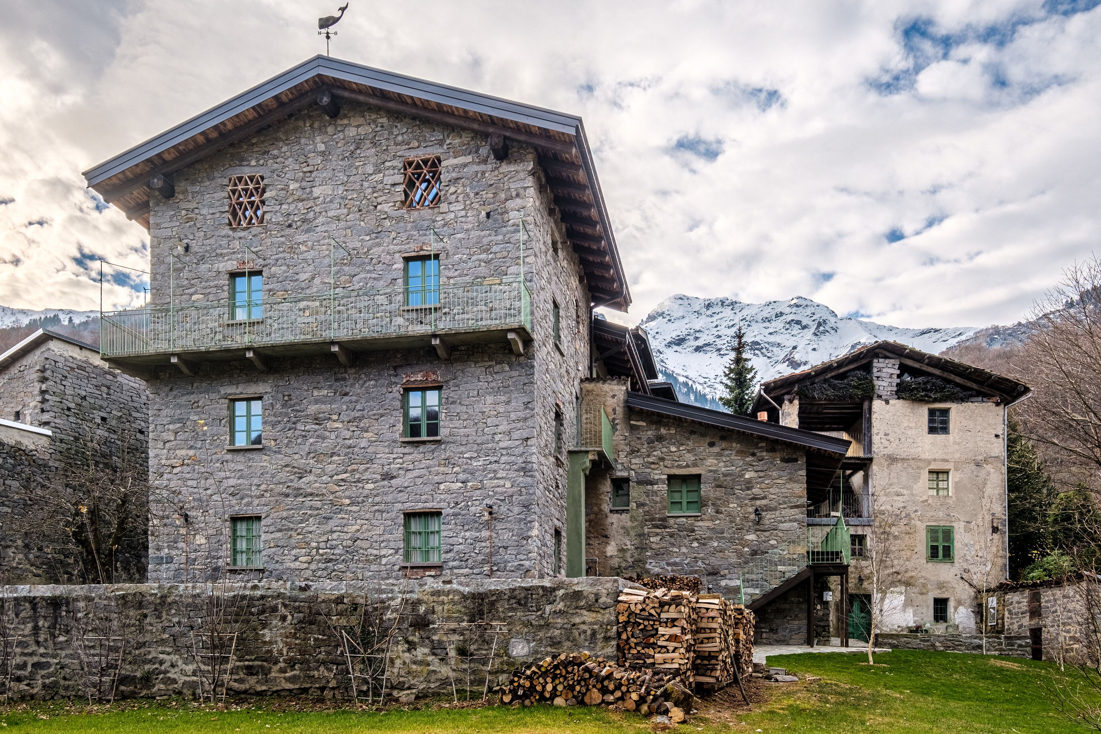 Un edificio de piedra con múltiples ventanas y balcones, rodeado de un área verde y montañas al fondo.