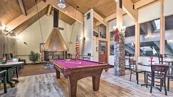 A pool table in a room with wooden floors and high ceilings, a stone pillar, and a view through large windows.