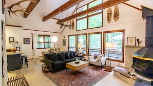 A spacious living room with a large sofa, a coffee table, and a fireplace. There are multiple windows providing natural light and a view of the outdoors. - Classic Cabin in Pocono Mountain Resort (Gouldsboro)