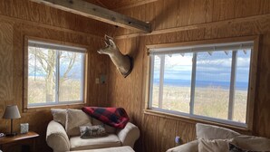 Una habitación acogedora con paredes de madera, una cabeza de ciervo montada en la pared, un sofá con una manta y ventanas amplias que ofrecen vistas del paisaje.