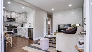 A modern kitchen with white cabinetry and stainless steel appliances, a dining area with a round table and chairs, and a living room with a sofa and a television.