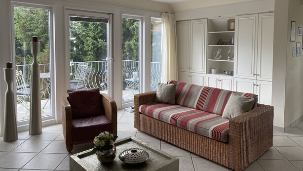 A spacious living area with a wicker sofa, a red armchair, and a coffee table with a vase of flowers. Large windows offer a view of the outdoors. - Villa Senator-Marstall - stylish and close to the beach (Travemünde)