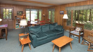 A cozy living room with a green sofa, wooden coffee table, and a dining table with chairs. There is a bookshelf with books and a lamp. Large windows offer a view of the forest. - The Keith Farm Cabin, right on peaceful BIG Lake -48 acres!!! (Presque Isle)