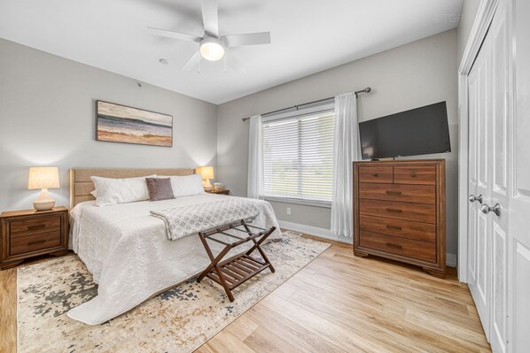 Une chambre à coucher comprenant un lit, des lampes de chevet, un ventilateur de plafond, un téléviseur posé sur un meuble, une commode en bois et une fenêtre avec des stores.