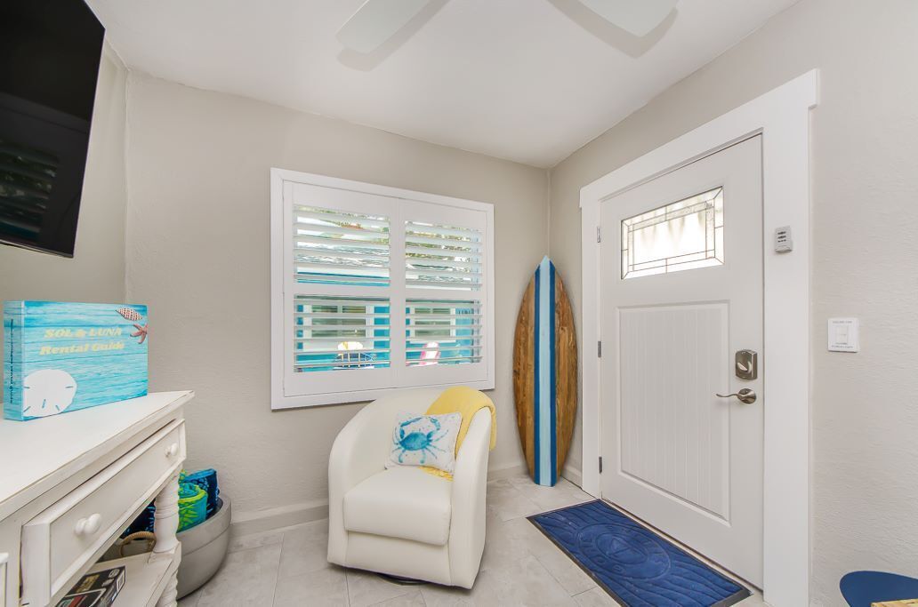 A room with a white door, a window with blinds, a white chair, a surfboard, and a box on a dresser.