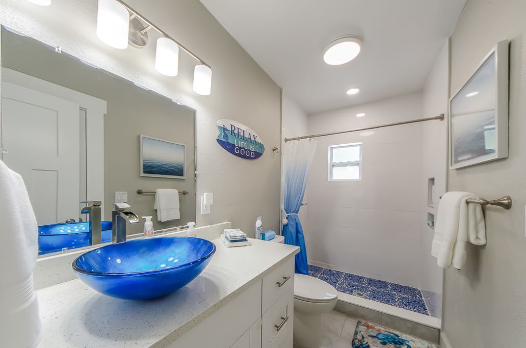 A bathroom with a large blue glass sink, a white toilet, a shower with a curtain, and a sign that says "RELAX LIFE IS GOOD".