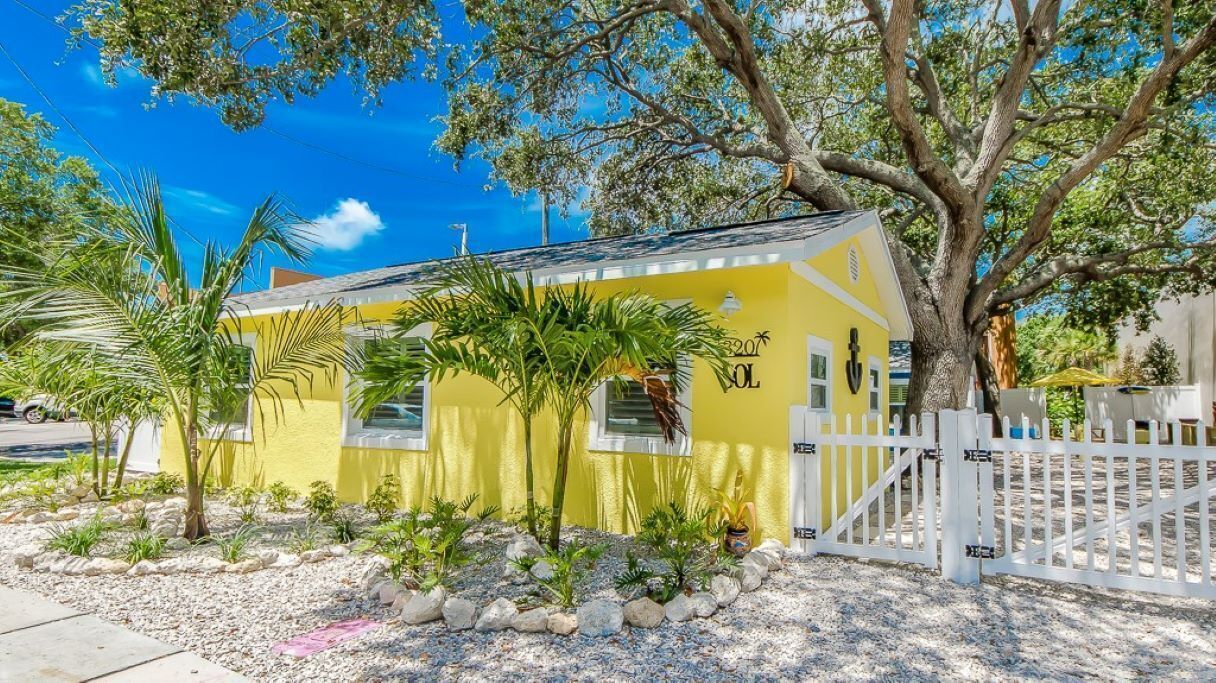 A yellow house with a white fence, surrounded by small plants and a large tree.