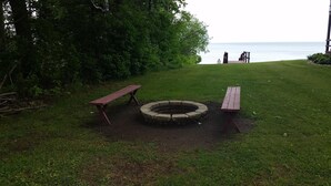 Un espacio con césped, una hoguera circular, dos bancos de madera y vistas al mar.