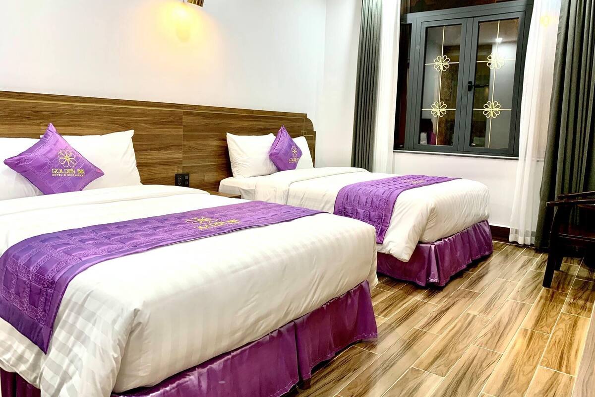 A hotel room with two beds, each with a purple bedspread and matching pillows, a wooden headboard, a nightstand, and a window with curtains.