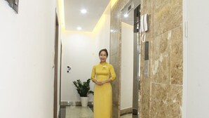 Mulher de vestido amarelo está em um corredor com paredes de mármore e um vaso de planta.