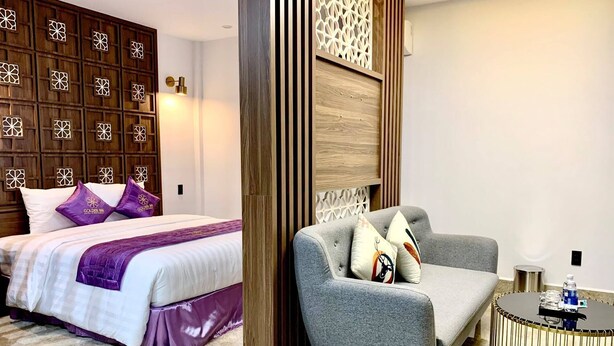 Quarto de hotel com cama, sofá cinzento, mesa de centro redonda e painel decorativo na parede.