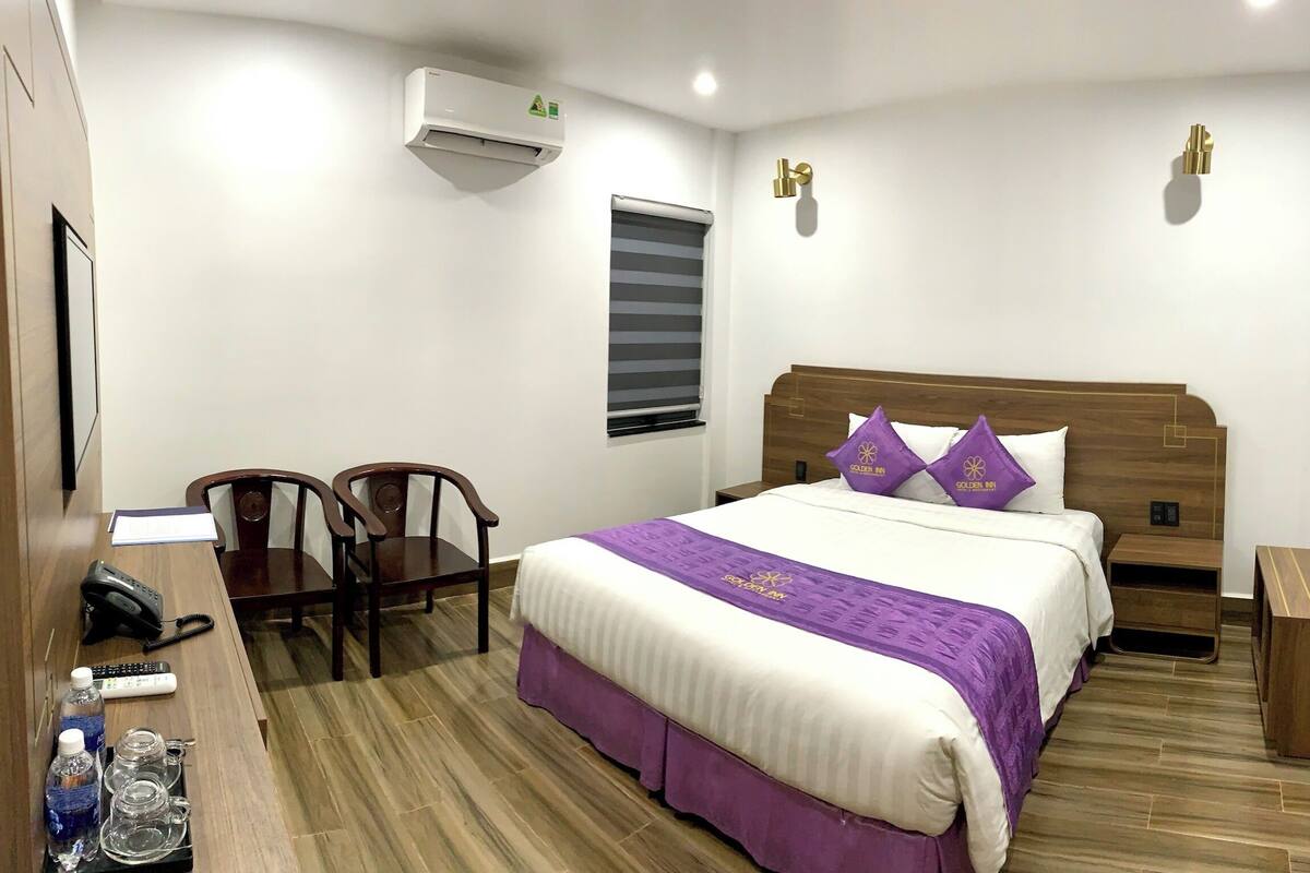 A hotel room with a wooden bed, a desk with a phone and remote controls, two chairs, and a window with blinds.
