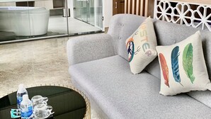 Uma sala de estar moderna com um sofá cinza, almofadas decorativas, uma mesa de centro redonda com garrafas de água e um box de chuveiro de vidro.