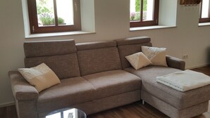 A modern living room with a sectional sofa, a glass coffee table, and a view of a garden through a window. - Vacation apartment in a farmhouse in beautiful Maxen in Müglitztal, near Dresden. (Müglitztal)