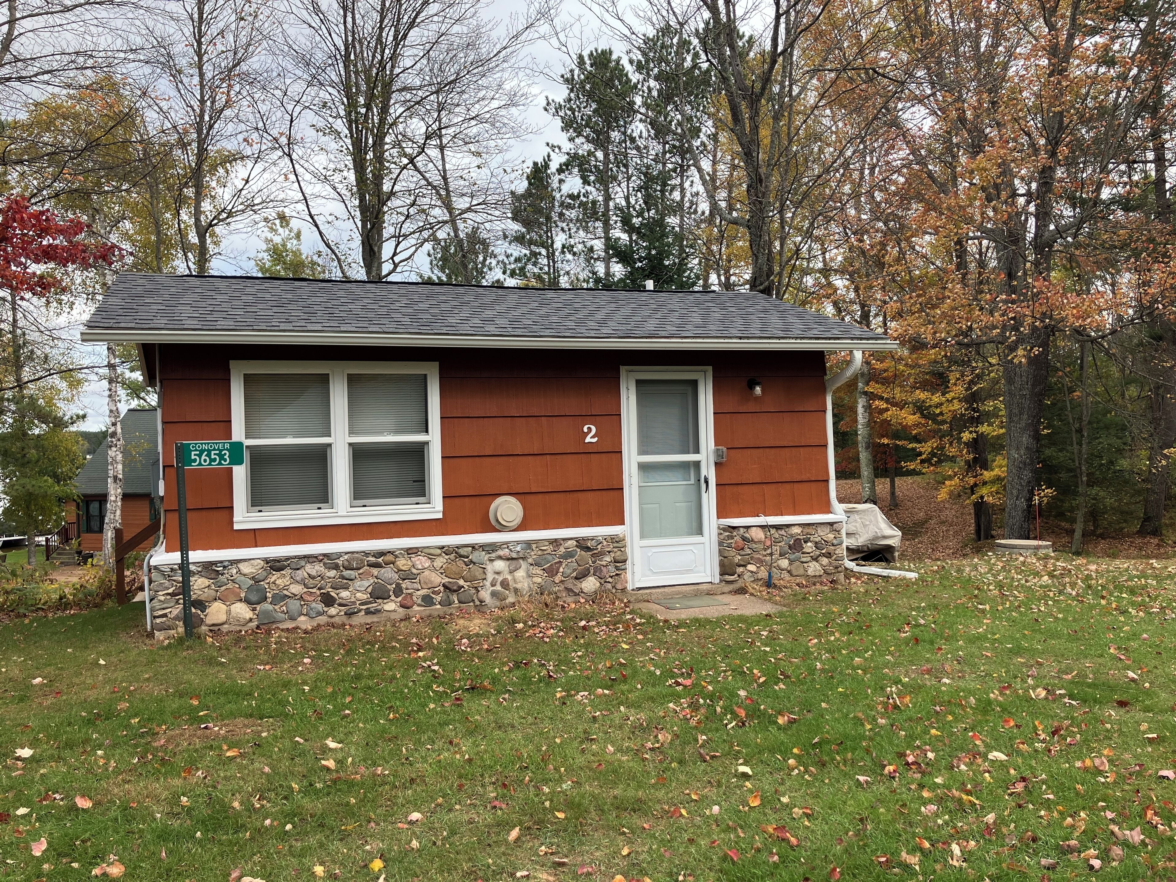 A small, single-story house with a stone foundation and a red exterior. It has a white front door, a window with a white frame, and a mailbox. The house is surrounded by a grassy yard with scattered fallen leaves.