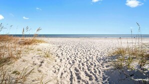 A sandy beach with footprints leading to the water, clear sky, and distant people. - Island Winds East 202 – Beachfront Condo Steps from The Hangout & Attractions (Gulf Shores)