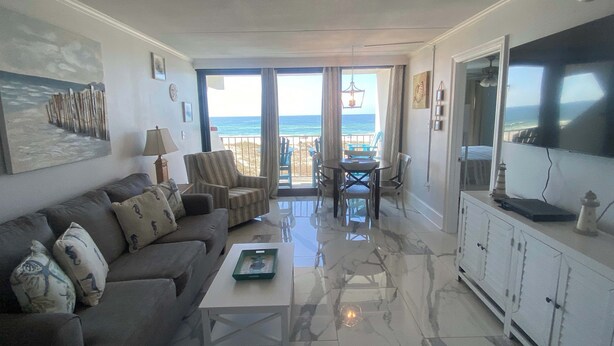 A living room with a sofa, coffee table, and a TV. There is a balcony with a view of the ocean. - Island Winds East 202 – Beachfront Condo Steps from The Hangout & Attractions (Gulf Shores)