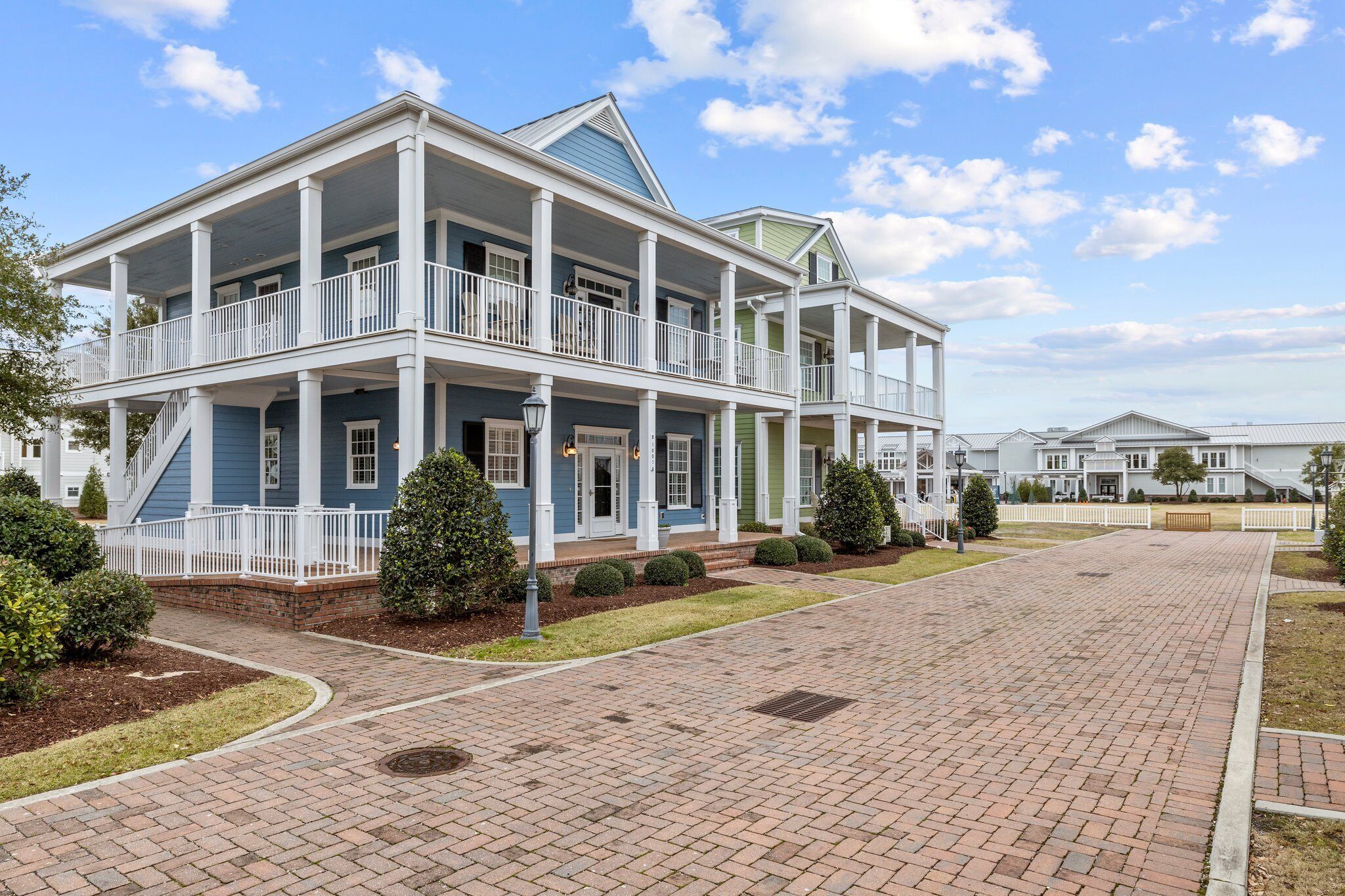 A two-story building with a covered porch and multiple balconies, surrounded by a brick pathway and small trees.
