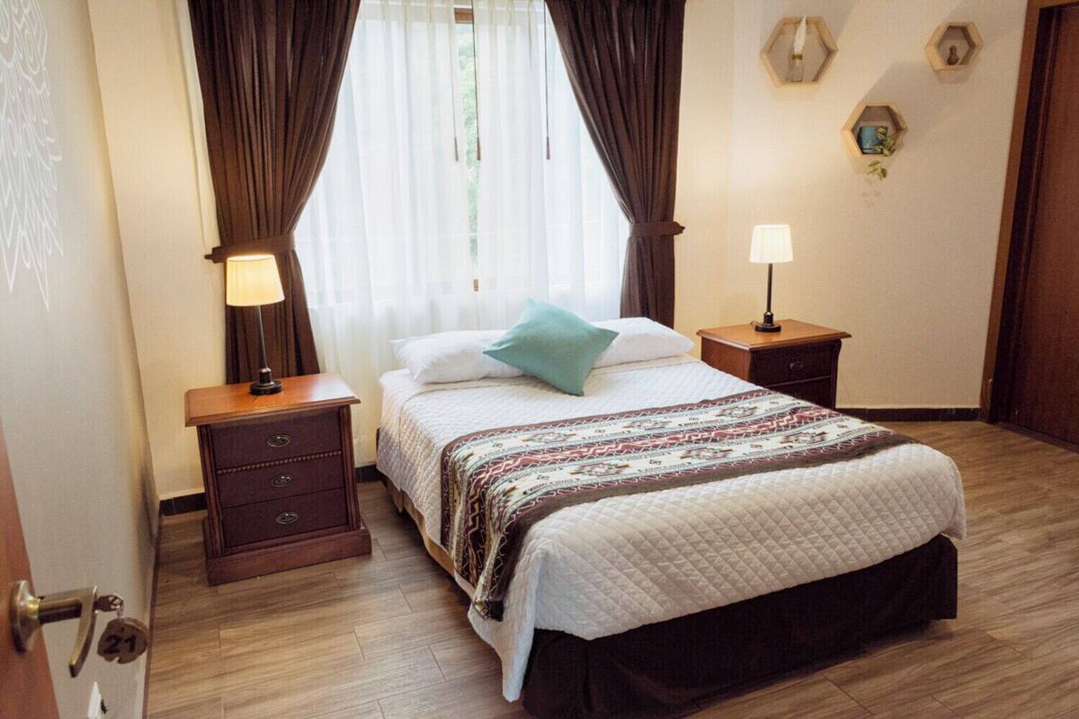 A hotel room with a bed, two bedside tables, a window with curtains, a wall-mounted lamp, and a door with a number.