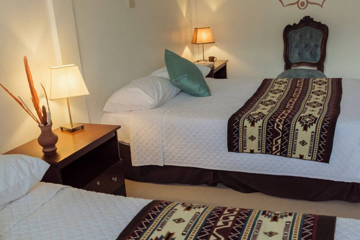 A hotel room with two beds, a decorative wall pattern, a nightstand with a lamp, and a hat on the bed.