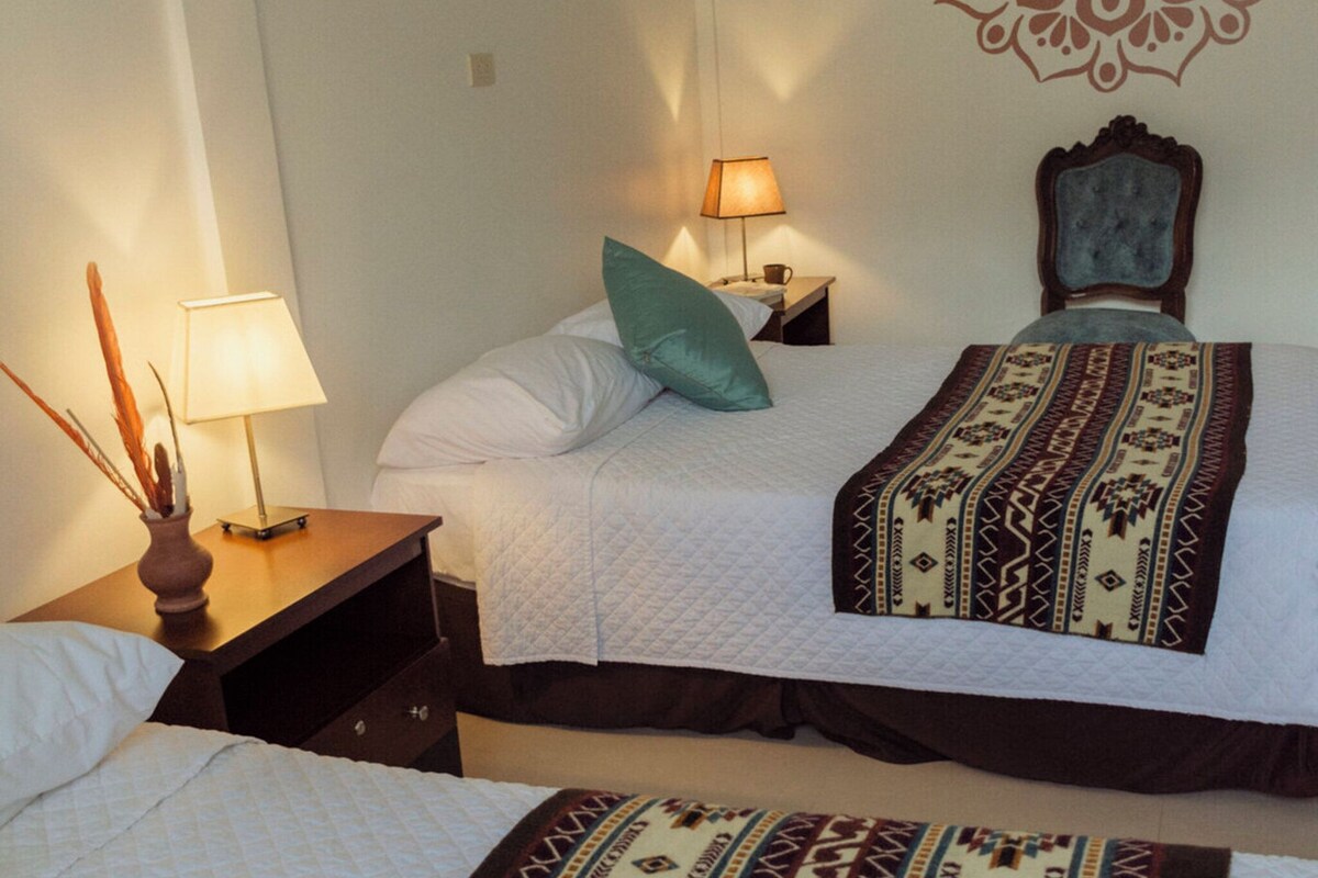 A hotel room with two beds, a decorative wall pattern, a nightstand with a lamp, and a window with blinds.