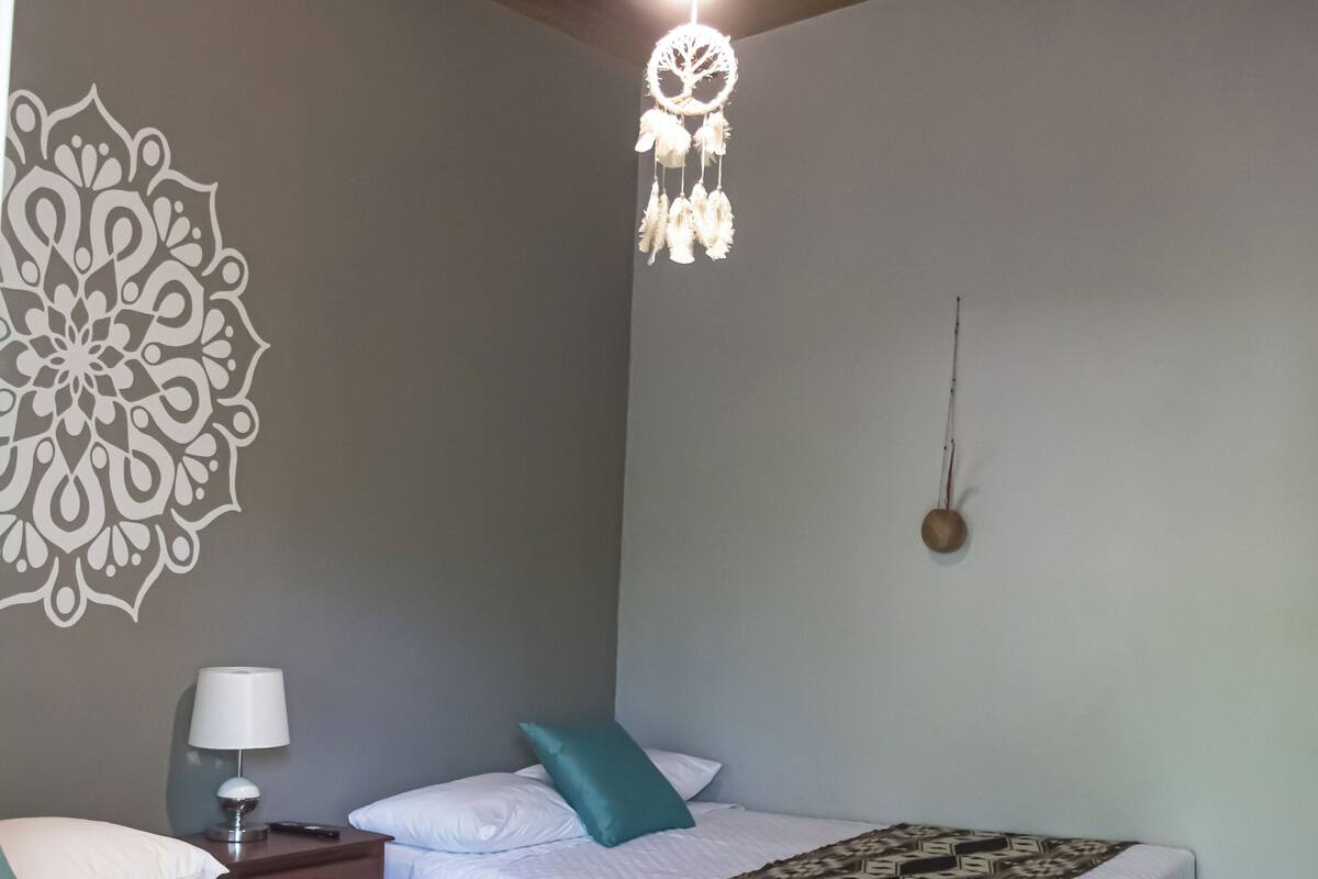 A bedroom with two beds, a nightstand, a lamp, a wall decoration, and a hanging light fixture.