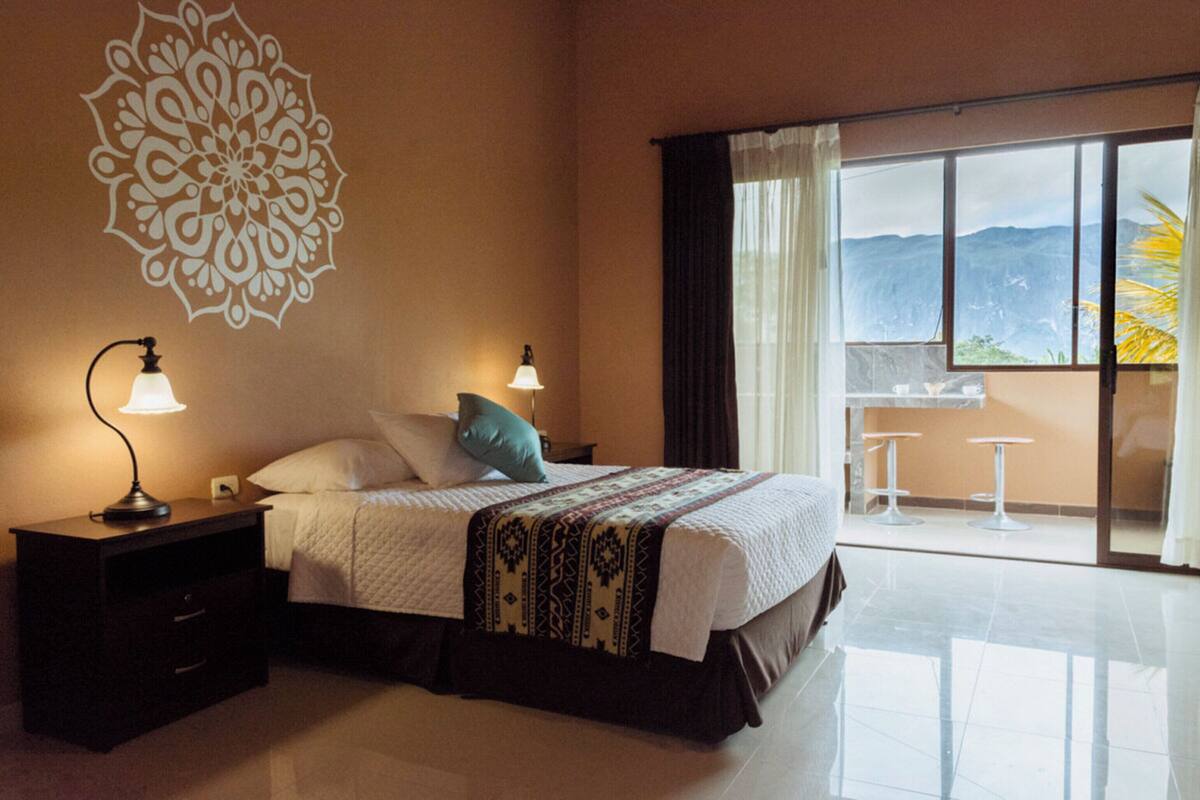 A hotel room with a bed, nightstands, a decorative wall piece, a balcony with a view, and a table with chairs.