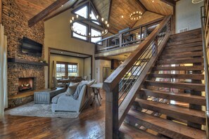 A spacious living area with a stone fireplace, a wooden staircase, and a high ceiling with exposed beams.