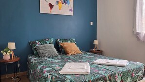 Een slaapkamer met een blauwe wand, een wereldkaart als kunstwerk, een bloemenbedekking, twee kussens, twee nachtkastjes met lampen, en een raam met gordijnen.