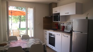 Een compacte keuken met witte kasten, een magnetron, oven en een koelkast. Er is een eettafel met stoelen en een patio paraplu zichtbaar door de schuifdeur.