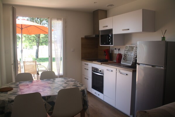 Eine kompakte Küche mit weißen Schränken, einer Mikrowelle, einem Backofen und einem Kühlschrank. Ein Esstisch mit Stühlen und ein Sonnenschirm sind durch die Glastür vom Balkon aus sichtbar.