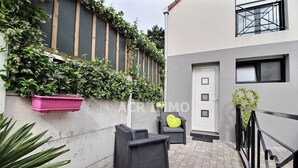 Un área de estar exterior moderna con una mesa y sillas de ratán negro, una maceta rosa y una puerta con ventanal.