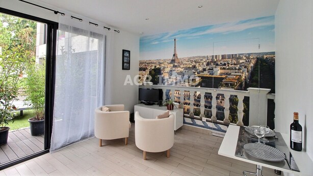 Una sala moderna con un ventanal grande, dos sillones blancos, un televisor de pantalla plana, una mesa de comedor con una botella de vino y un mural de un paisaje urbano en la pared.