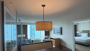 A hotel room with a dining area, a large window with a view of buildings, a bed, and a ceiling lamp. - Luxury Beachfront Resort 1BR Suite w/ Full Kitchen in VA Beach Sleeps 4+ (Virginia Beach)