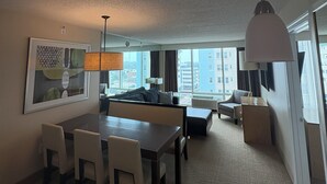 A hotel room with a dining area, a sofa, a chair, a table, and a large window with a city view. - Luxury Beachfront Resort 1BR Suite w/ Full Kitchen in VA Beach Sleeps 4+ (Virginia Beach)