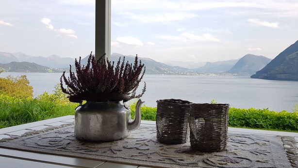 A view of a lake with mountains in the background, a metal teapot, and two wicker baskets on a table. - Irenegarden (Volda)