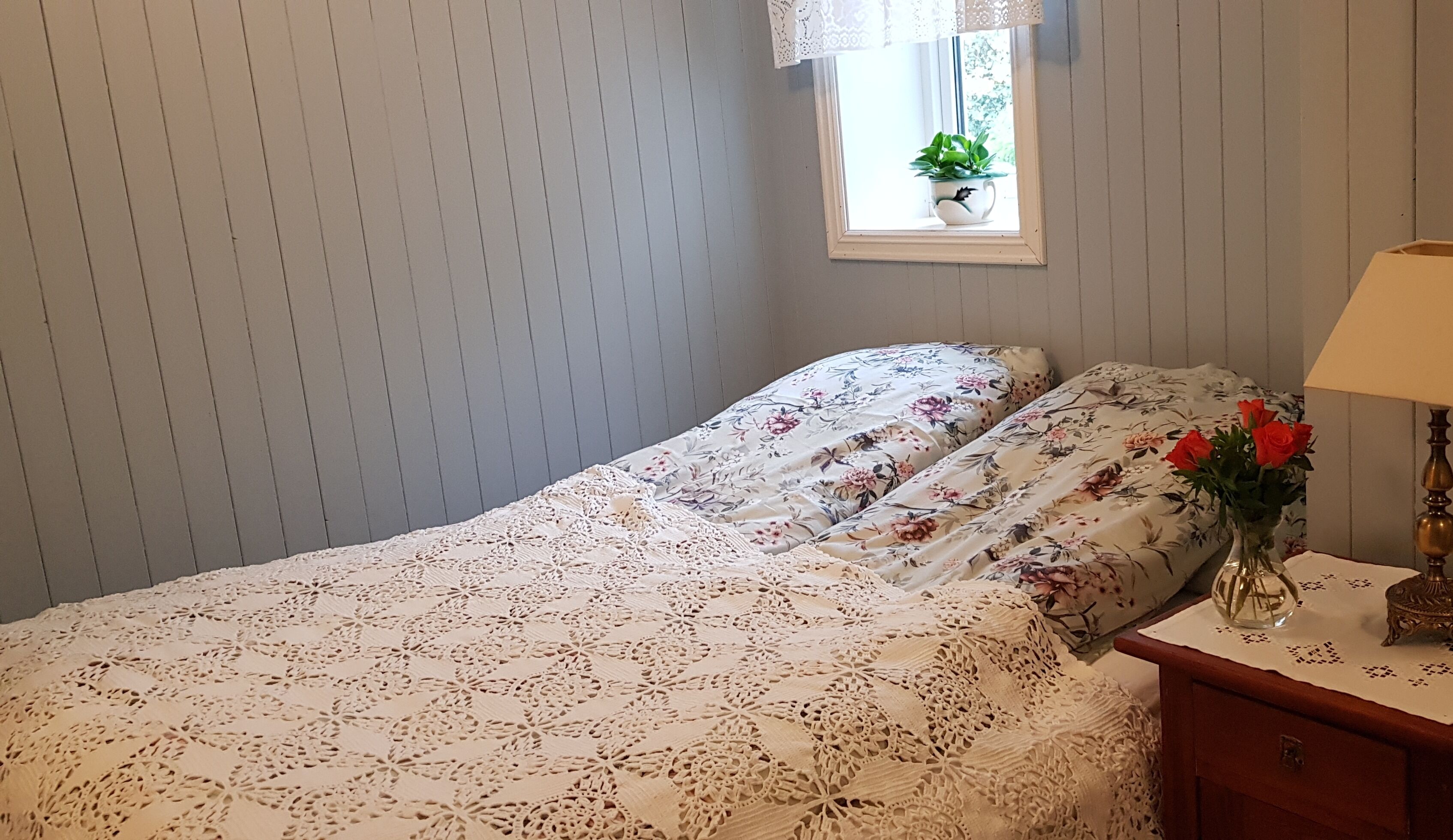 A bedroom with a bed covered in floral bedding, a lace doily, a nightstand with a lamp, and a window with a plant.