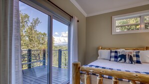 Ein Schlafzimmer mit einer großen Schiebeglastür, die auf eine Außenterrasse führt, einem Bett mit karierter Bettwäsche und einem Fenster mit Blick auf Bäume.