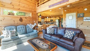 L’intérieur chaleureux d’un chalet comprend un espace de vie avec un canapé bleu et un divan en cuir marron, un plafond en bois et une cuisine visible à travers une porte ouverte.