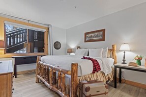 Un dormitorio con estructura de cama de madera, un escritorio, una silla, una ventana con vistas y una mesita de noche con lámpara.