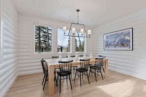 Uma sala de jantar com mesa e cadeiras de madeira, janelas amplas e um lustre.