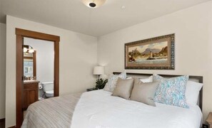 Relax in a spacious room with a large bed, adorned with a white comforter and decorative pillows, and a beautiful coastal scene above.