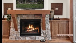 Una chimenea con detalles de piedra, un televisor de pantalla plana montado encima y una vista de una casa al fondo.