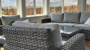 Ein Zimmer mit grauem Sofa, Korbstühlen und Blick auf einen Holzzaun und Hügel.
