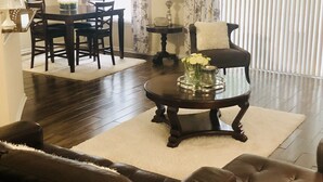 A living room with a dark wood floor, a brown leather sofa, a coffee table with a vase of flowers, a dining area with a table and chairs, and a window with blinds. - Beautiful home with pool (Orange Park)