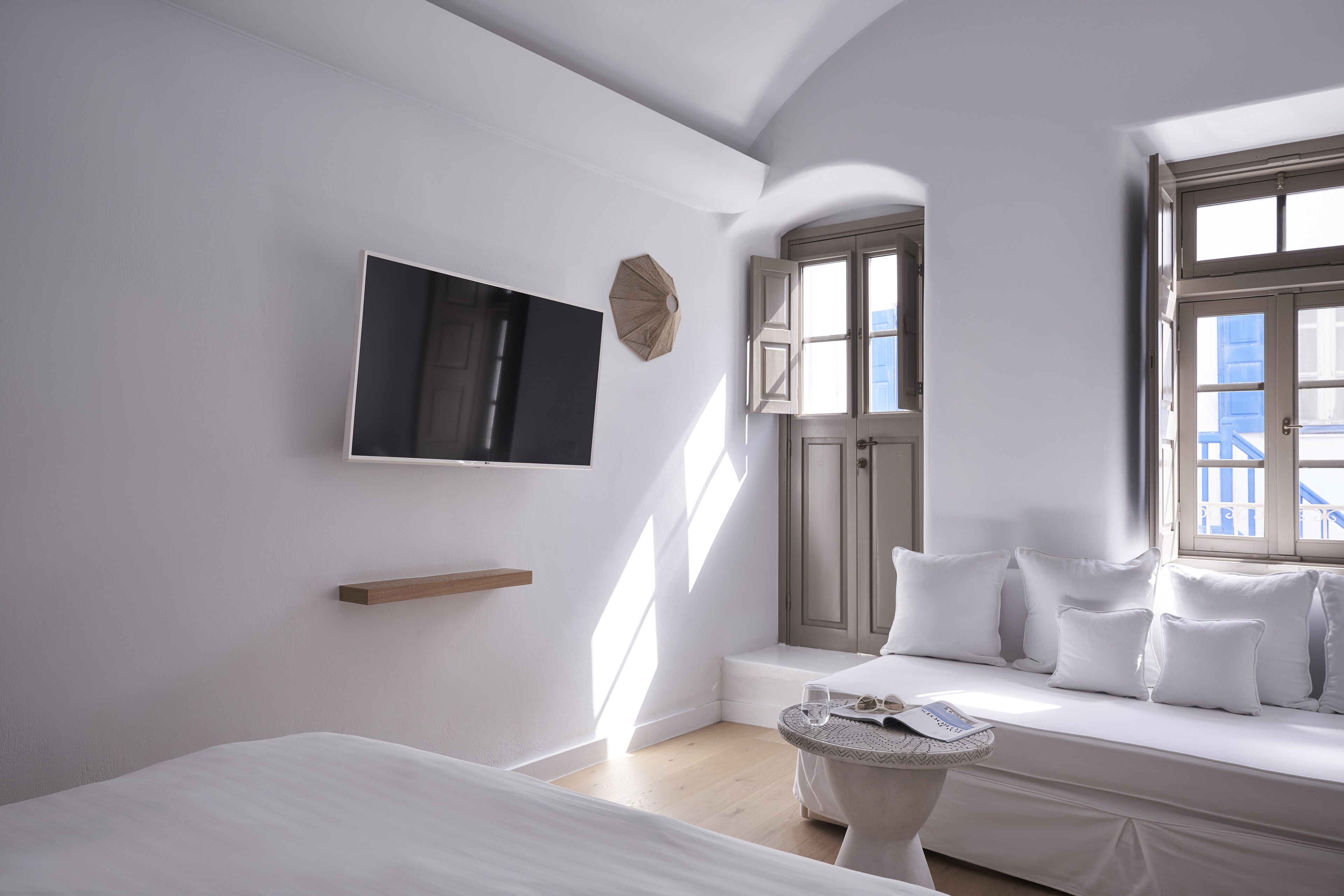 A modern hotel room with a bed, a seating area with cushions, a television mounted on the wall, and a window with a view of the outside.