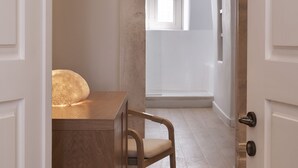 Un couloir étroit mène à une pièce dotée d’une grande fenêtre, d’une chaise en bois et d’une petite table avec une lampe.